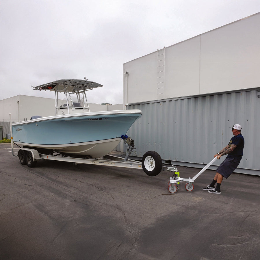 Person using a manual Trailer-Tug 3.0 trailer dolly to move a tandem-axle boat trailer with a light blue boat across level concrete